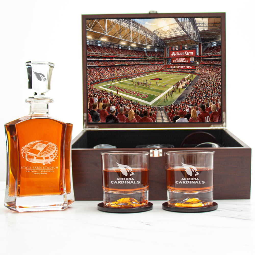 A whiskey gift set featuring a clear glass decanter filled with amber liquor, two matching engraved glass rocks tumblers with amber liquid and orange slices, and an open dark wood presentation box showing a printed stadium scene. The set is photographed on a light neutral background.