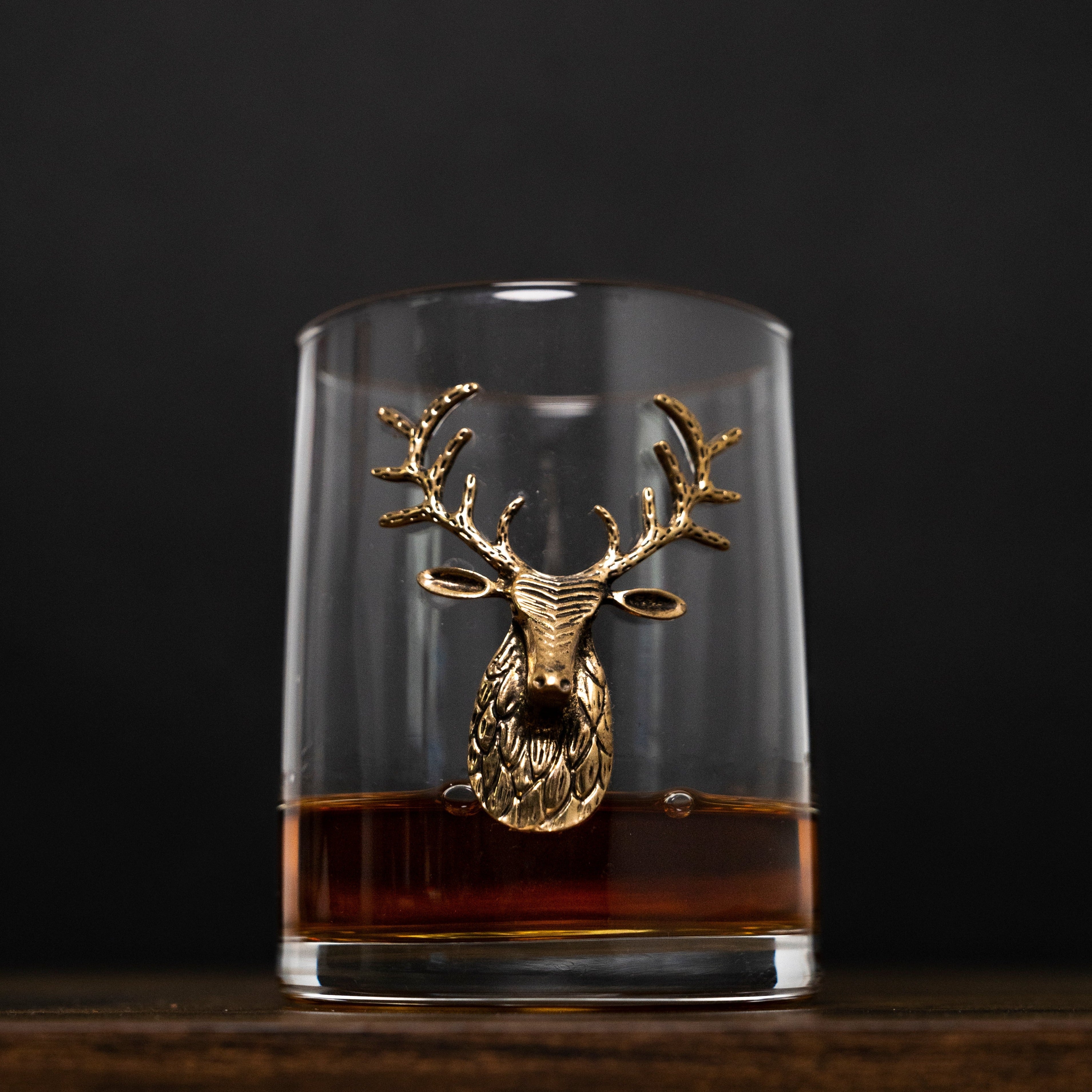 Glass tumbler with gold deer head design filled with a dark liquid on a black background