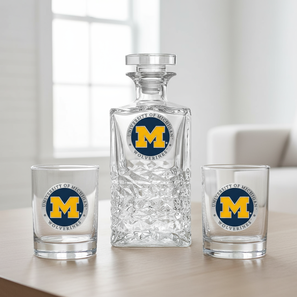 Clear glass decanter and two glasses with University of Michigan Wolverines logo on a light background